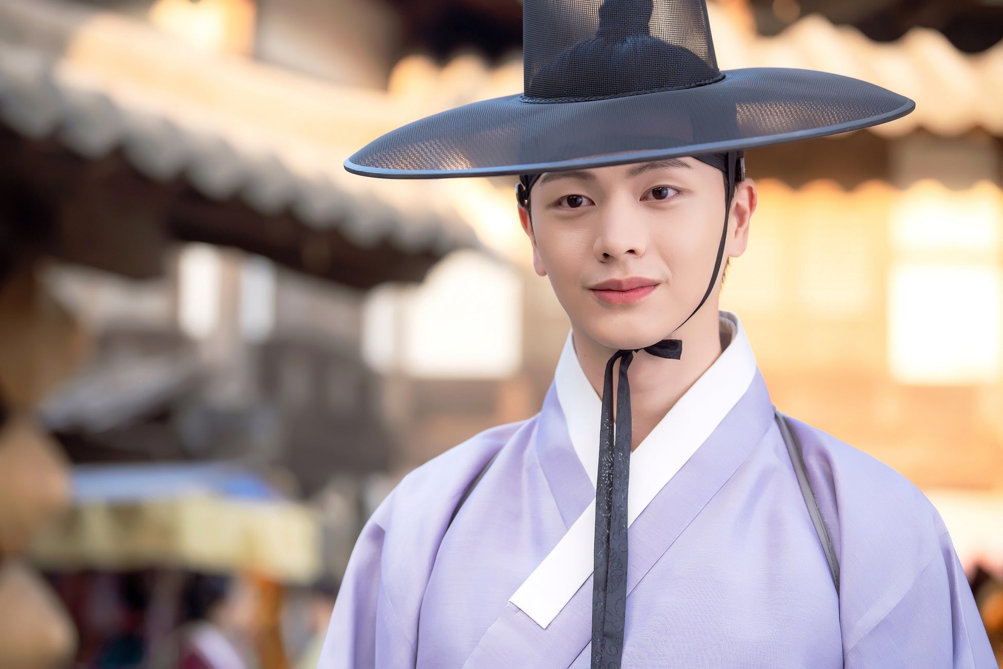 この画像は、韓国の時代劇に登場するキャラクターを演じる俳優の写真です。俳優は伝統的な韓国の衣装を着用しており、淡い紫色の上着と白い襟が特徴的です。頭には黒いガト(韓国の伝統的な帽子)をかぶっています。背景には、ぼんやりとした伝統的な建物の一部が見え、時代劇の雰囲気を醸し出しています。俳優は穏やかな表情をしており、全体的に落ち着いた雰囲気が漂っています。