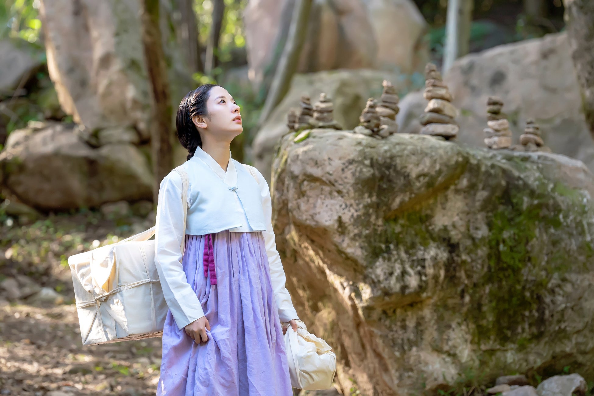 この画像は、韓国ドラマ「鬼宮(ききゅう)」の一場面を捉えたものです。女性が伝統的な韓服を着ており、白い上着と淡い紫色のスカートを身にまとっています。彼女は森の中に立ち、何かを見上げている様子です。背景には大きな岩があり、その上には小さな石が積み重ねられています。周囲は緑豊かな自然に囲まれており、静かな雰囲気が漂っています。このシーンは、ドラマのファンタジー要素や時代劇の雰囲気を強調しています。
