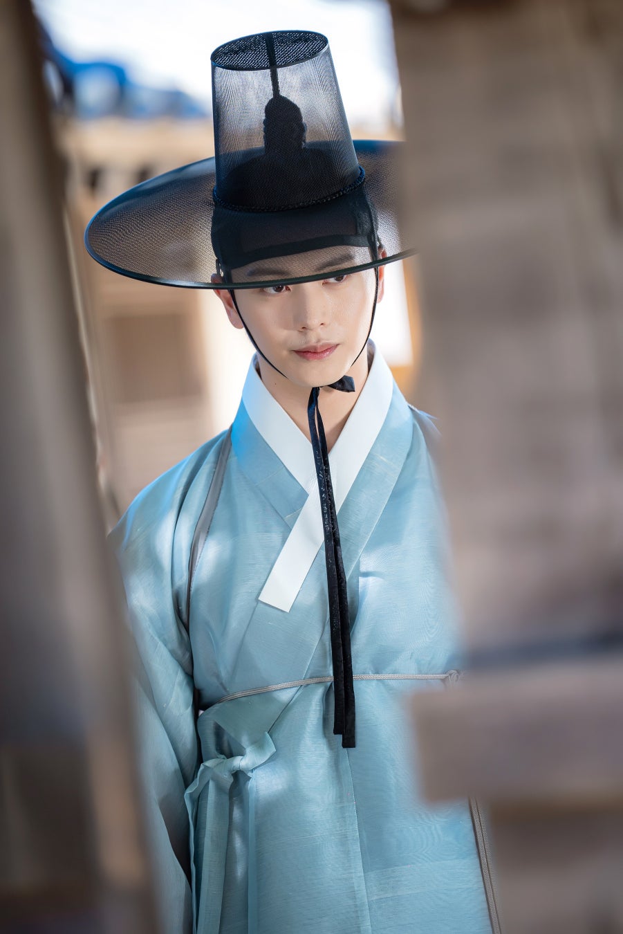 この画像には、伝統的な韓国の衣装を着た男性が写っています。彼は淡い青色のハンボクを身にまとい、黒いガト(韓国の伝統的な帽子)をかぶっています。背景はぼかされており、建物の一部が見えます。彼の表情は落ち着いており、視線はカメラの方を向いています。全体的に、時代劇の雰囲気を感じさせるシーンです。