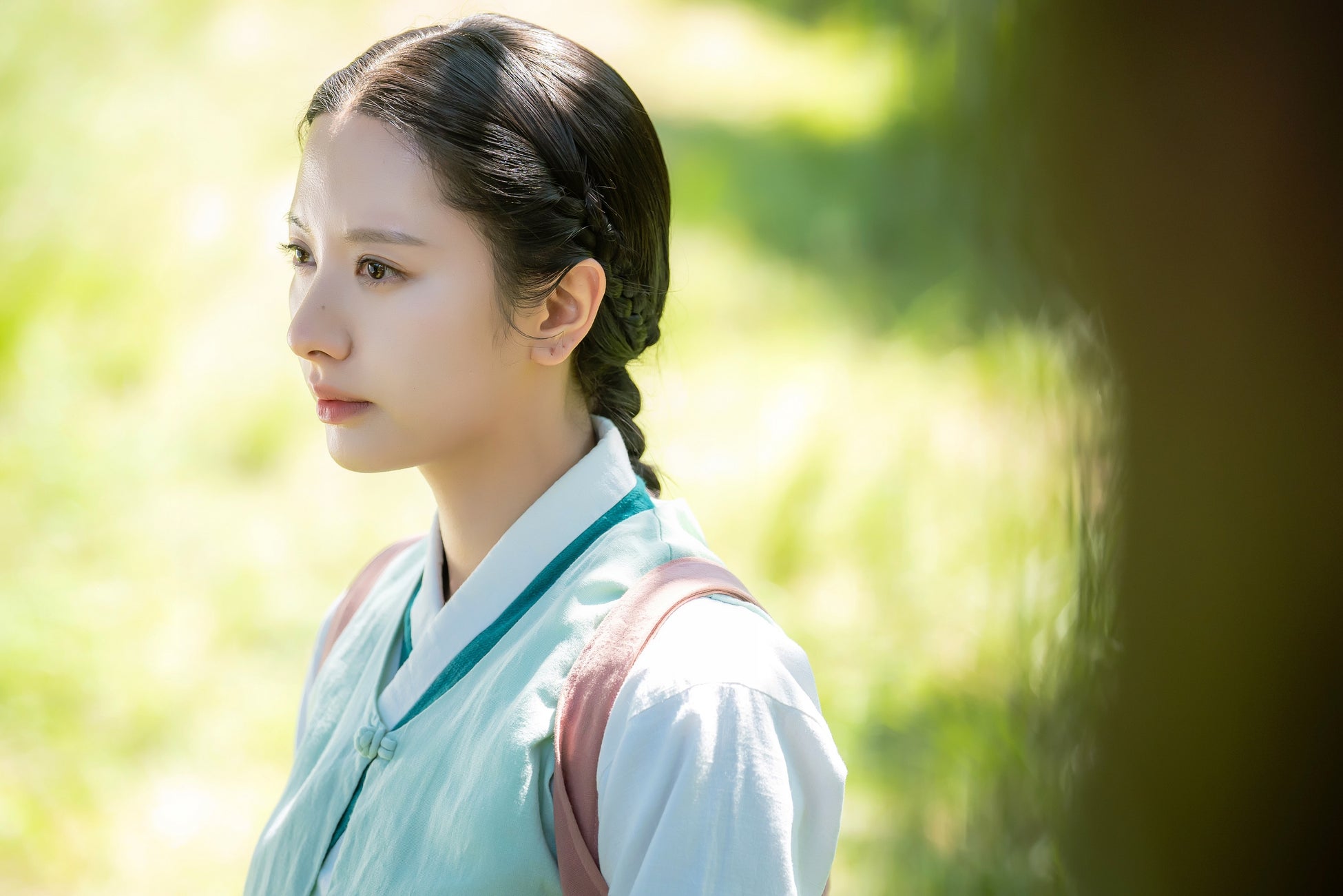 この画像は、韓国ドラマ「鬼宮(ききゅう)」のシーンを捉えたものです。女性が伝統的な韓服を着ており、背景には緑豊かな自然が広がっています。彼女は横を向いており、穏やかな表情をしています。髪はきちんと編み込まれており、全体的に落ち着いた雰囲気が漂っています。光が柔らかく差し込み、彼女の顔を優しく照らしています。
